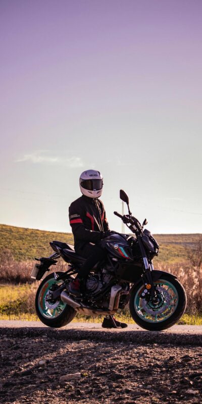 A motorcyclist in a leather jacket and helmet sits on a motorcycle on a rural road under a clear sky.