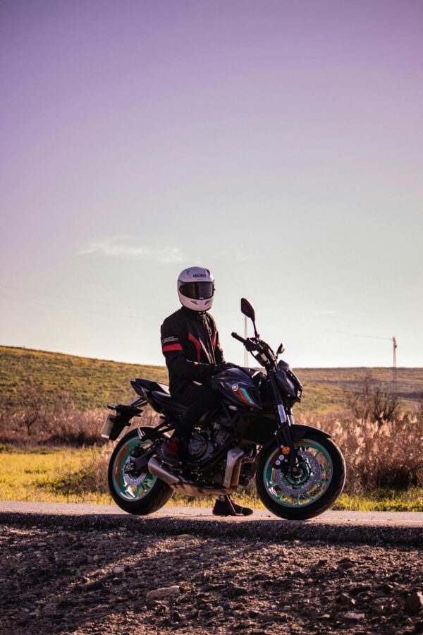 A motorcyclist in a leather jacket and helmet sits on a motorcycle on a rural road under a clear sky.