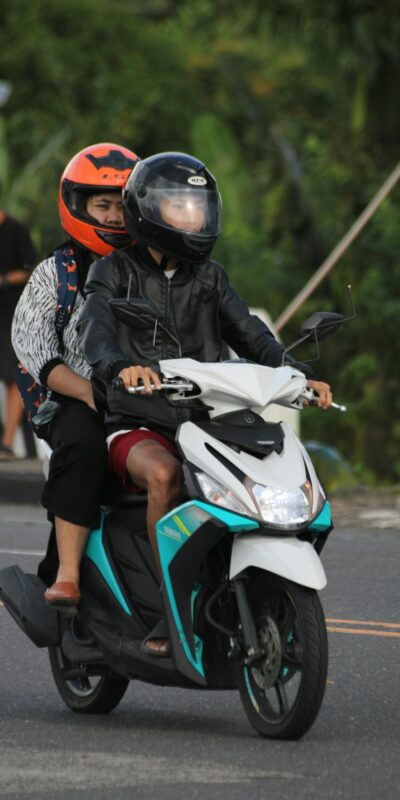 Two people on a scooter wearing helmets, riding on a city road.