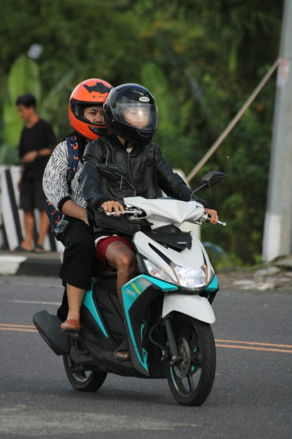 Two people on a scooter wearing helmets, riding on a city road.